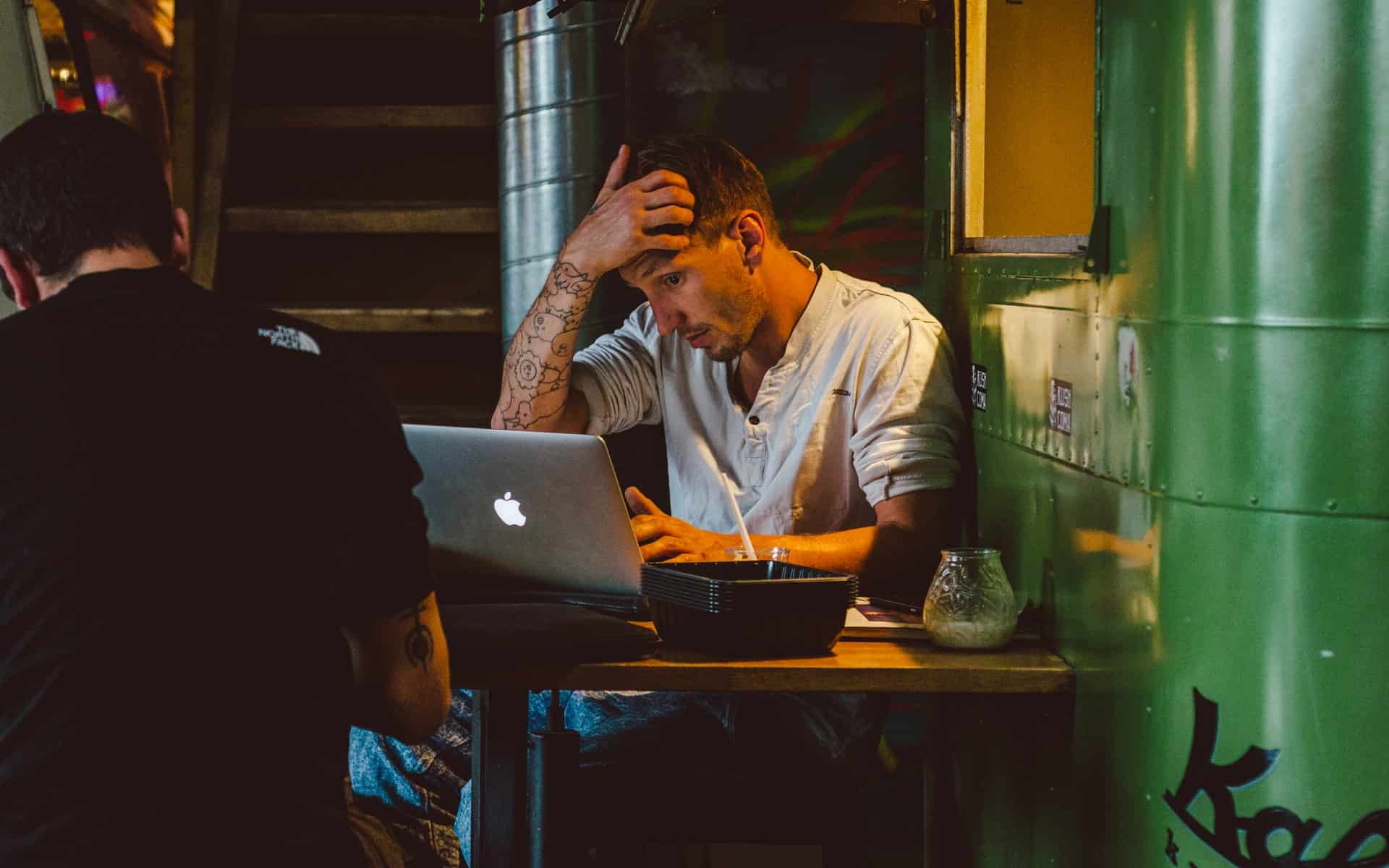Man with hand on head in front of laptop in cafe