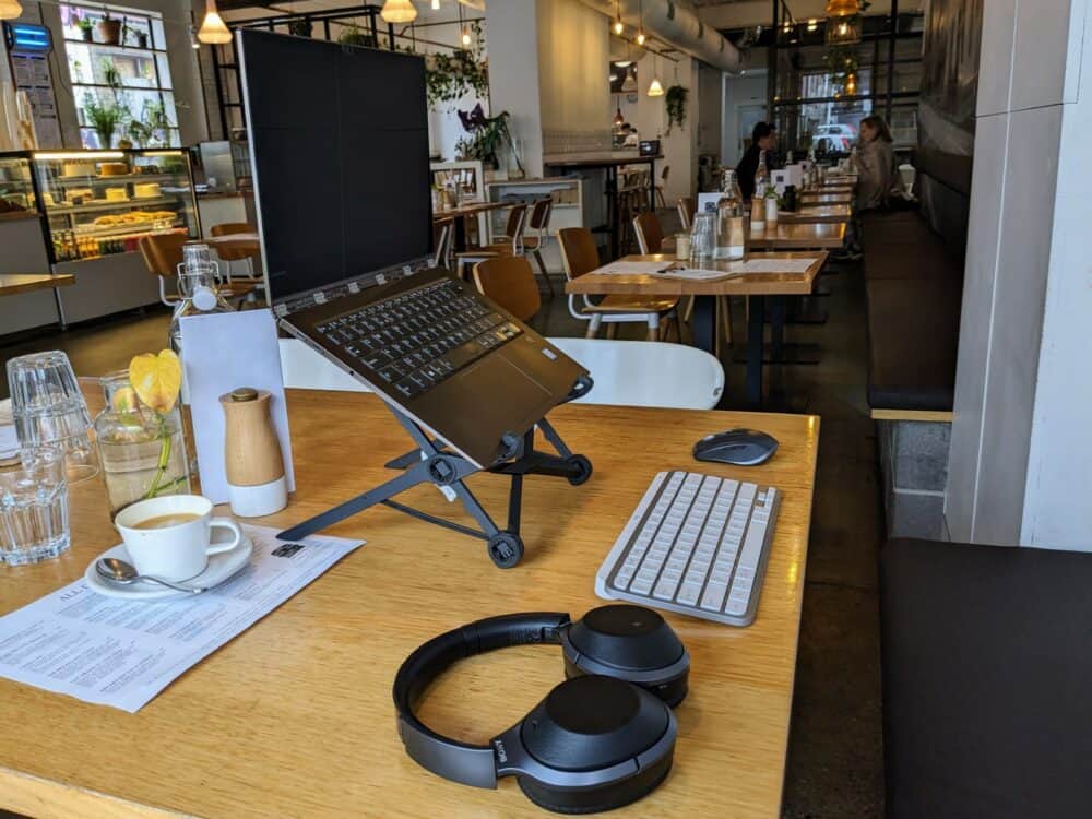 Portable office setup in a cafe, with a laptop on a stand, keyboard and mouse, and headphones on a wooden table
