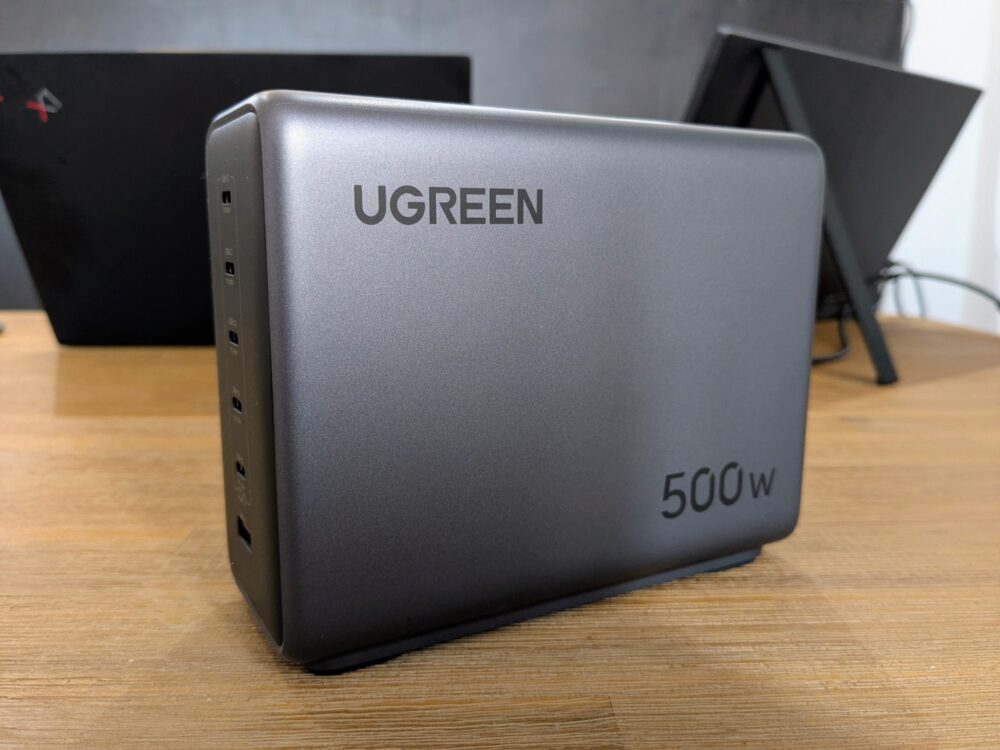 UGreen 500W charger sitting at an angle on a wooden table with a laptop and portable monitor behind
