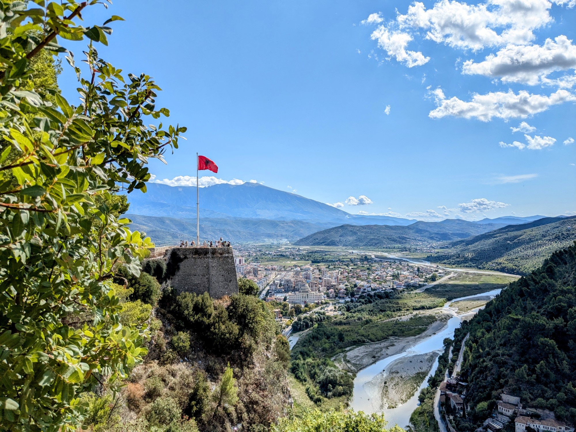 Expansive view from alongside a hilltop castle, looking towards a stone tower with a large flag on it. Below lies a river, with a town along both banks.