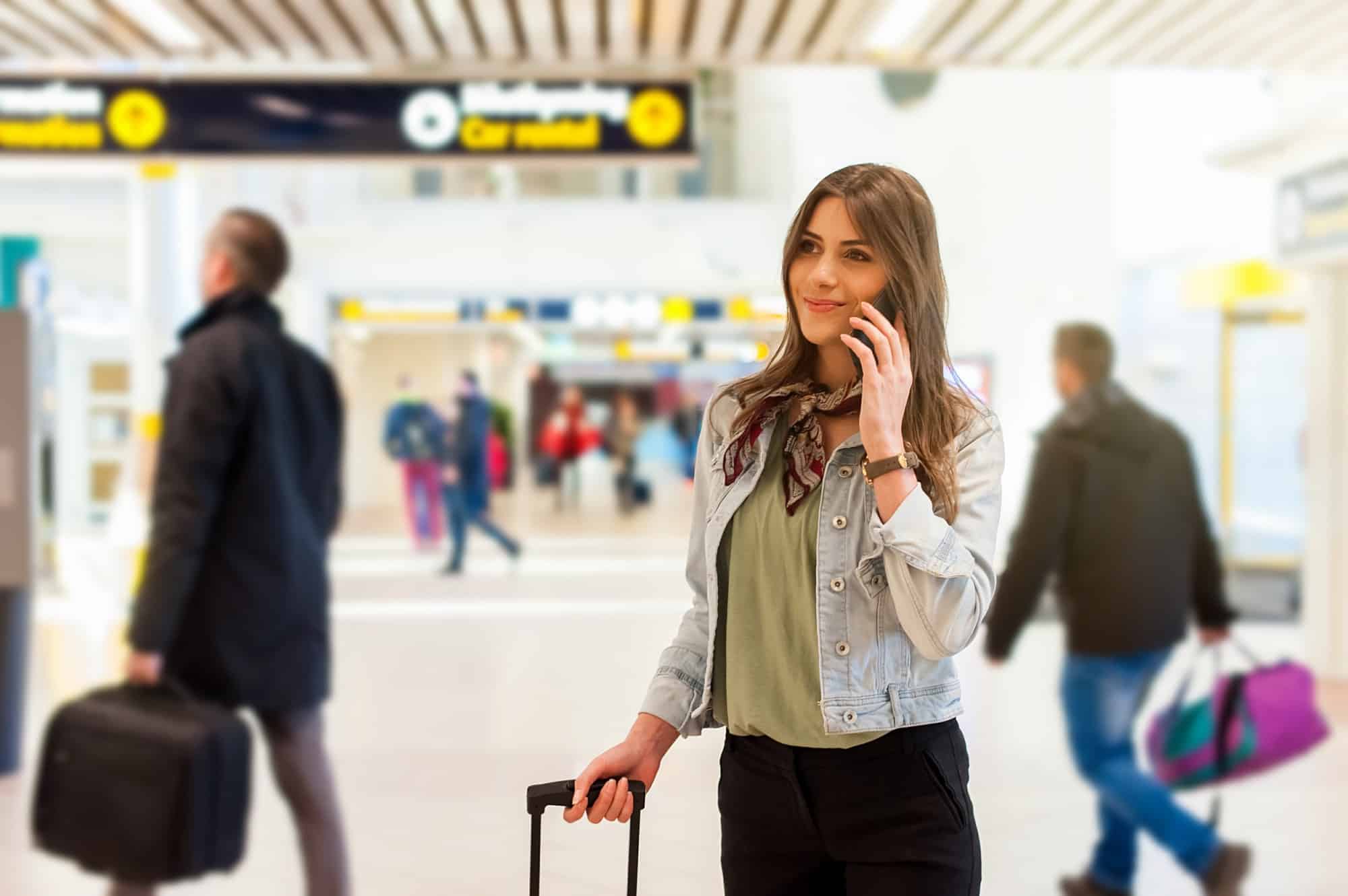 Young woman talking on a smartphone at an airport, holding the handle of a suitcase with people walking in the background