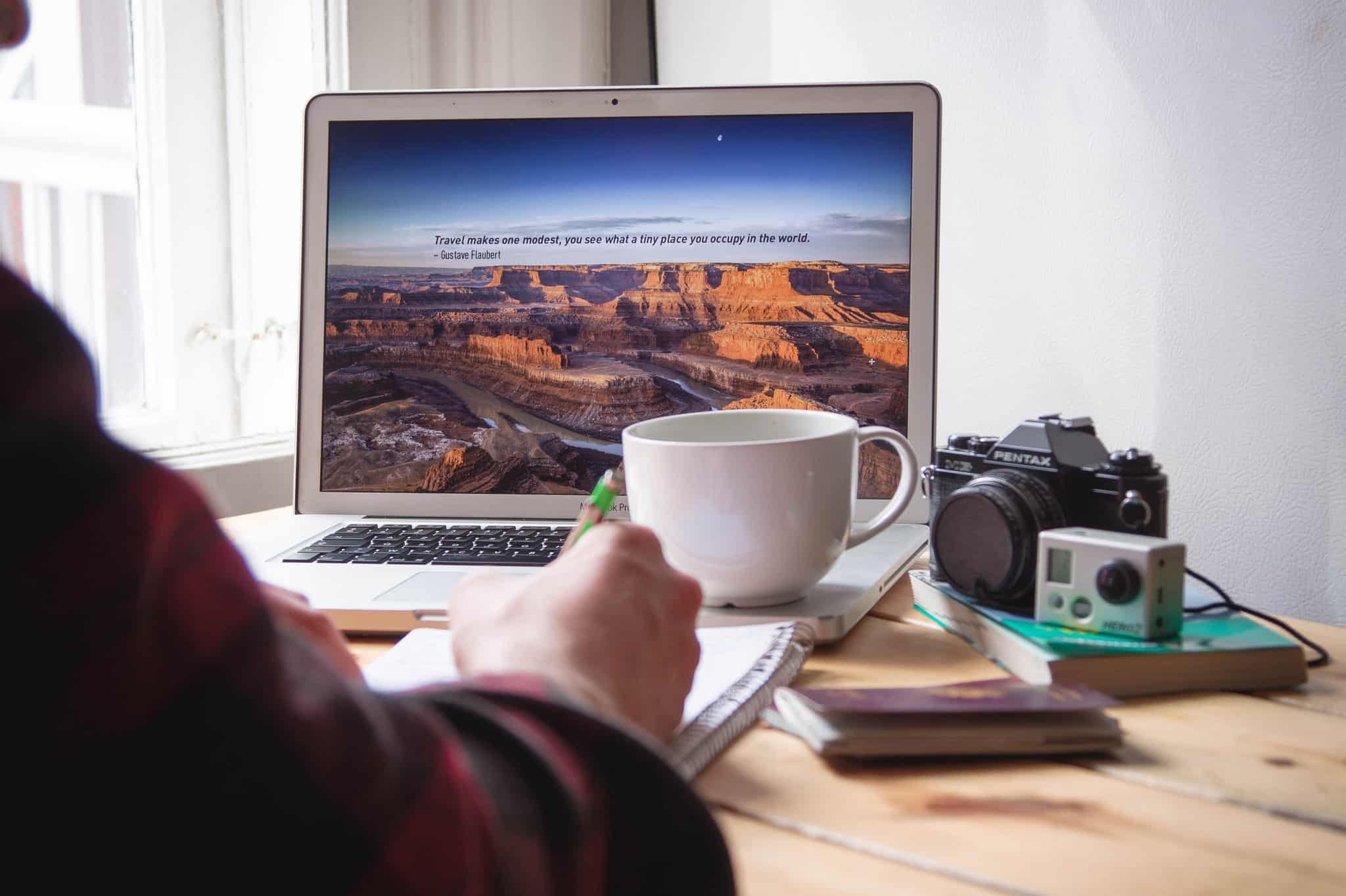 Arm of person writing in a notepad with a laptop and coffee cup on a wooden table beyond, along with a book, DSLR, and action camera.