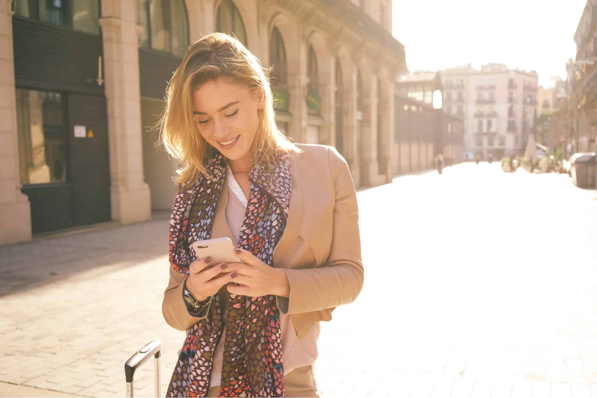 Young woman with suitcase checking phone on city street